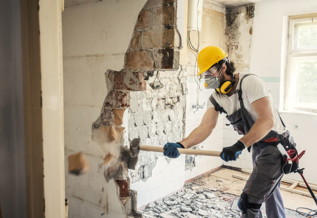 Contractor wearing a respirator and safety gear demolishing a wall during a home renovation as part of a spring checklist for asbestos awareness.