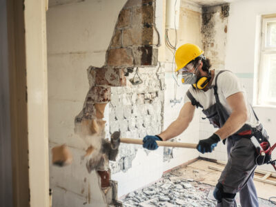 Contractor wearing a respirator and safety gear demolishing a wall during a home renovation as part of a spring checklist for asbestos awareness.